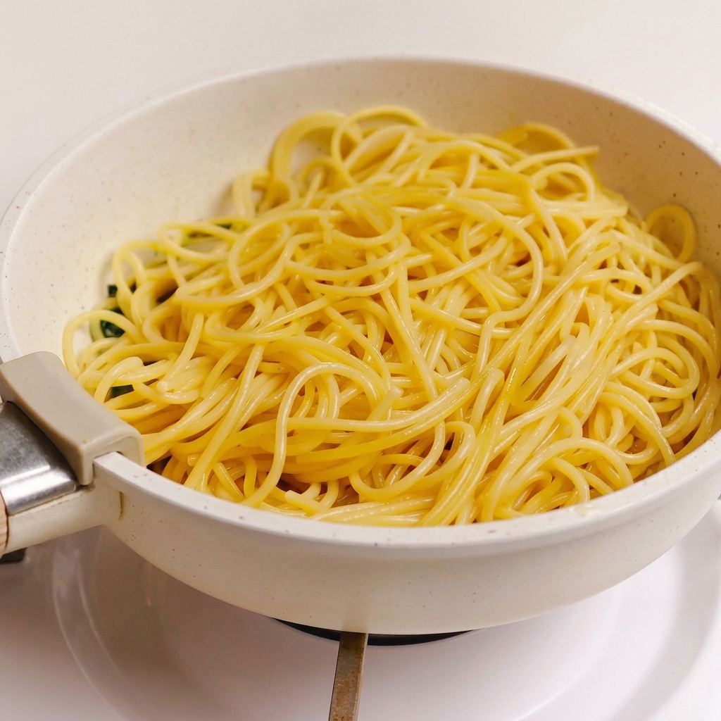 Ugotowany makaron spaghetti wyłożony na sos z zielonych ziół, czosnku i boczku na jasnej patelni.
