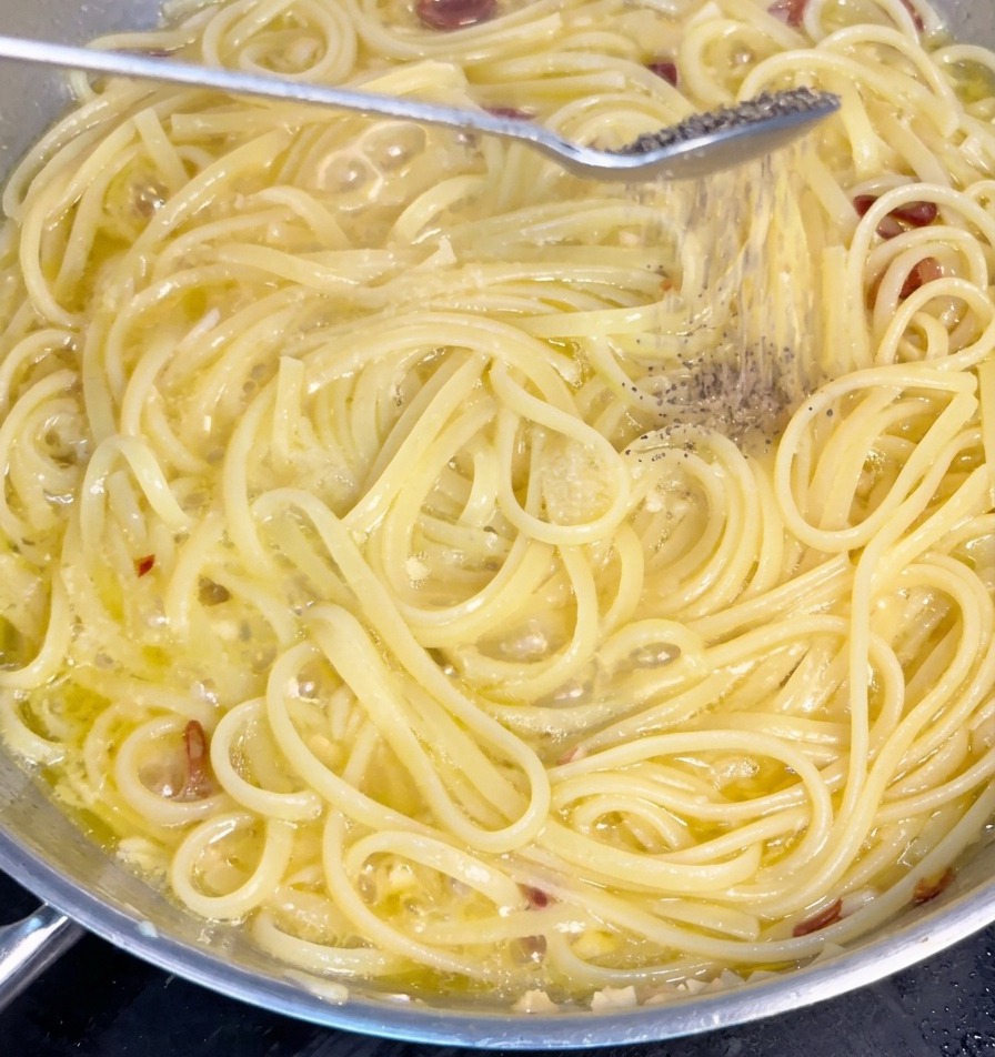 Świeżo mielony czarny pieprz sypiący się na makaron spaghetti na patelni z sosem czosnkowo-oliwnym.