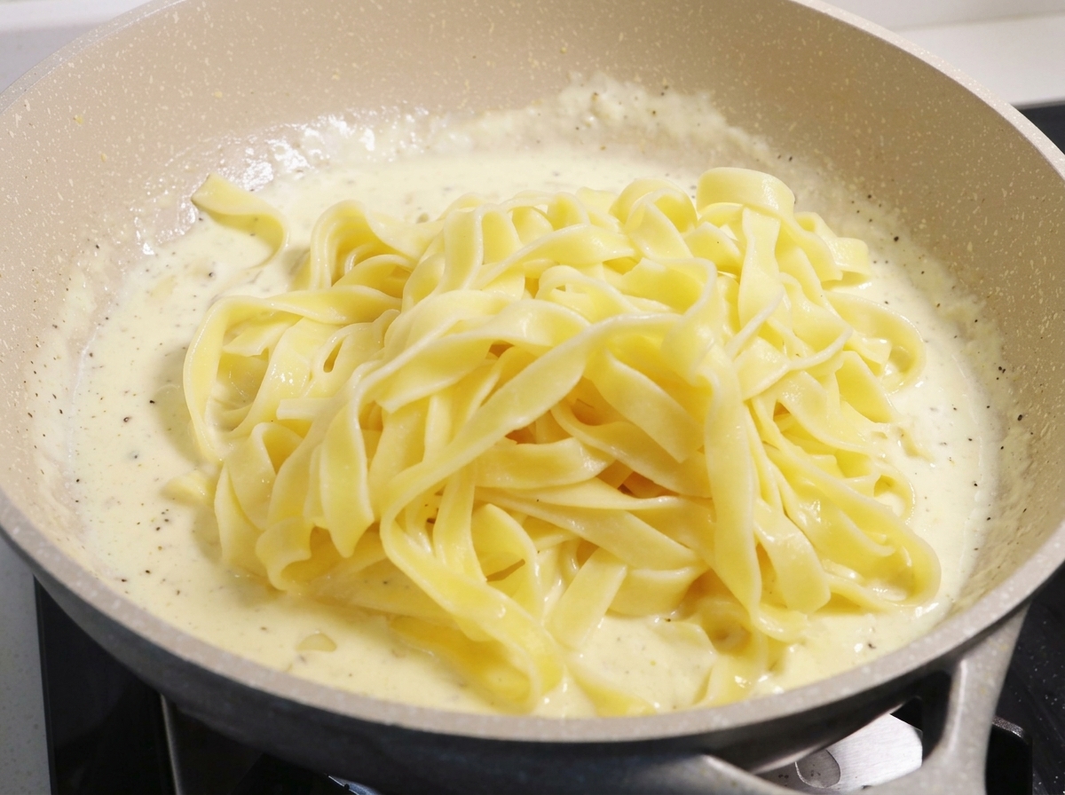 Duża porcja ugotowanego żółtego makaronu fettuccine ułożona na gęstym, bulgoczącym białym sosie śmietanowym na dużej patelni.