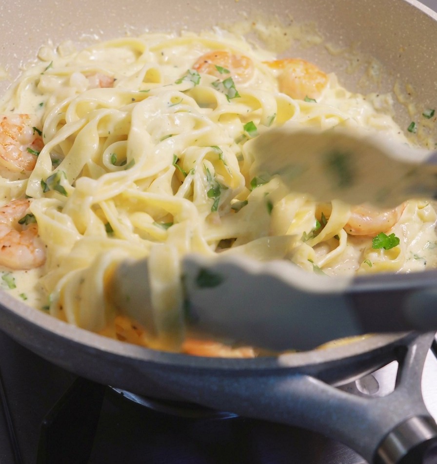 Używanie szczypiec do podnoszenia i mieszania makaronu fettuccine oraz różowych krewetek w kremowym białym sosie na nakrapianej patelni.