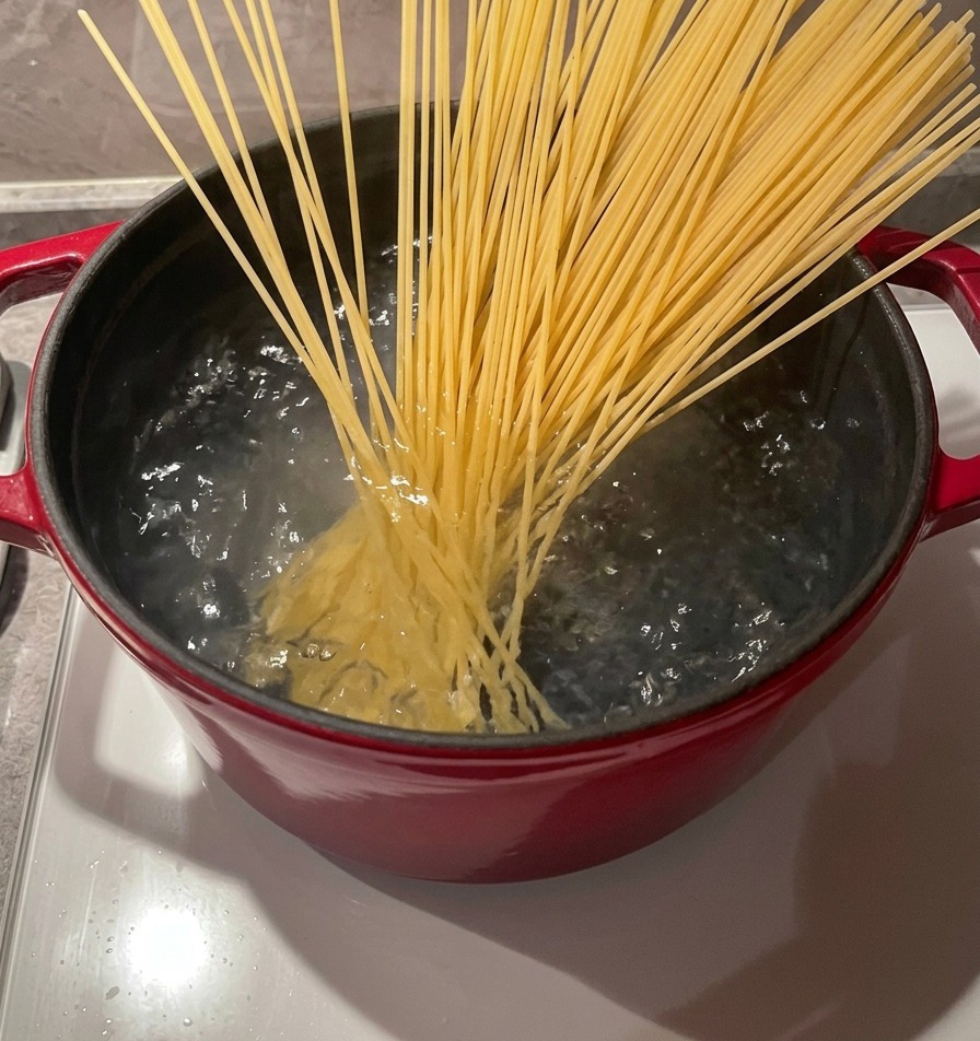 Pęczek suchego makaronu spaghetti wkładany za pomocą drewnianych pałeczek do garnka z bulgoczącą, wrzącą wodą.
