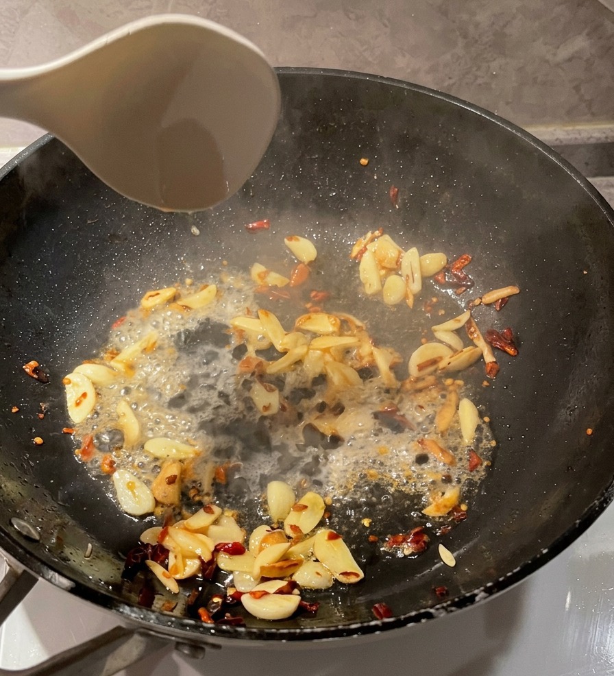 Biała chochelka wlewająca klarowną wodę z makaronu na czarną patelnię z podsmażonym czosnkiem i płatkami chili.