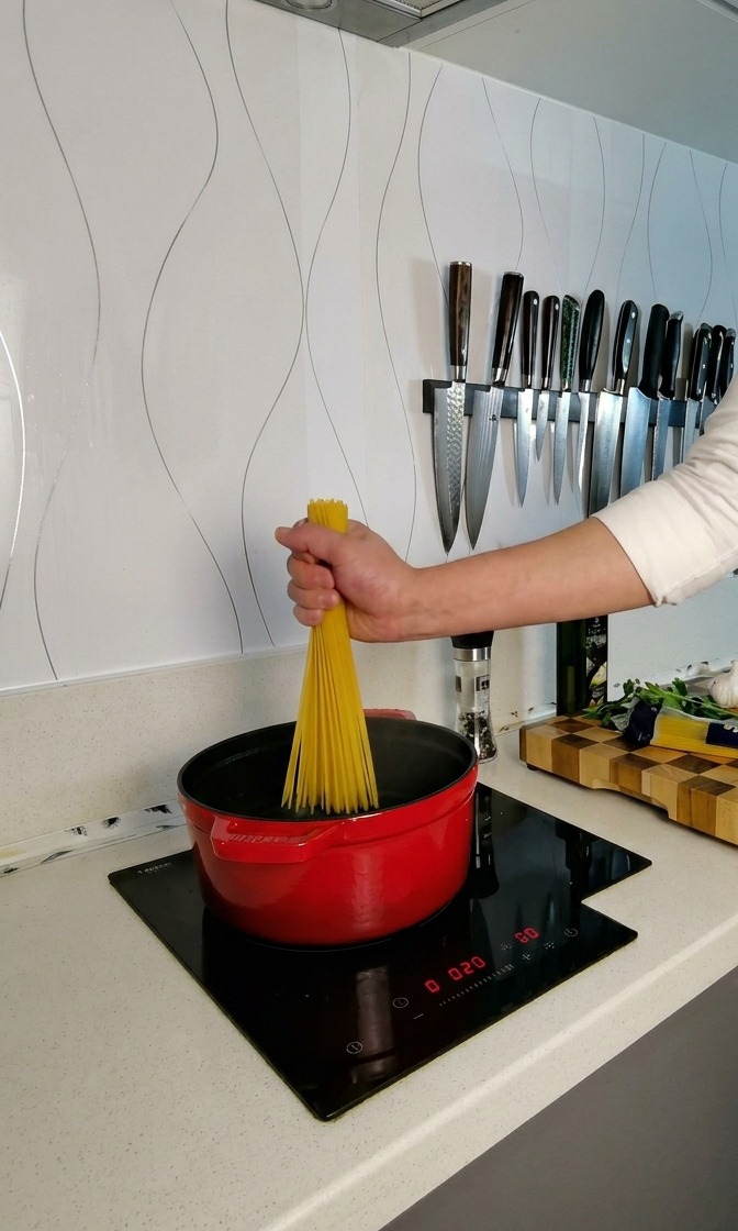 Dłoń trzymająca pęk suchego spaghetti nad czerwonym garnkiem z wrzącą wodą, skręcająca makaron tak, by rozłożył się w wachlarz.