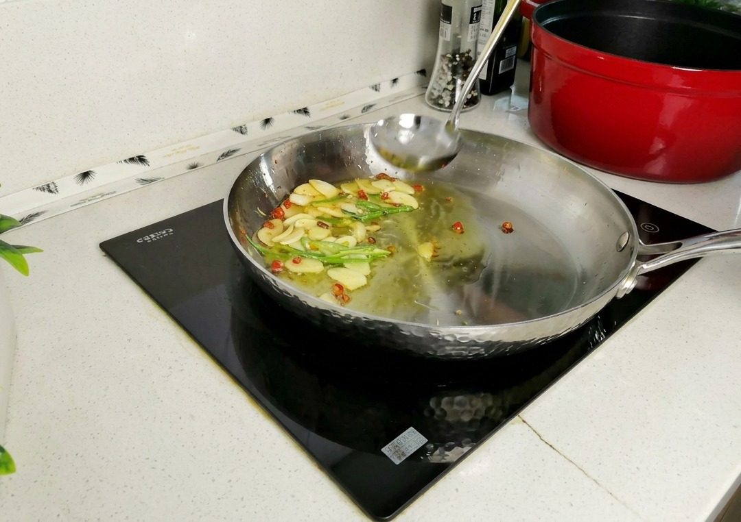 Chochla wlewająca mętną wodę po makaronie na patelnię ze smażonymi plasterkami czosnku, chili i łodygami pietruszki.
