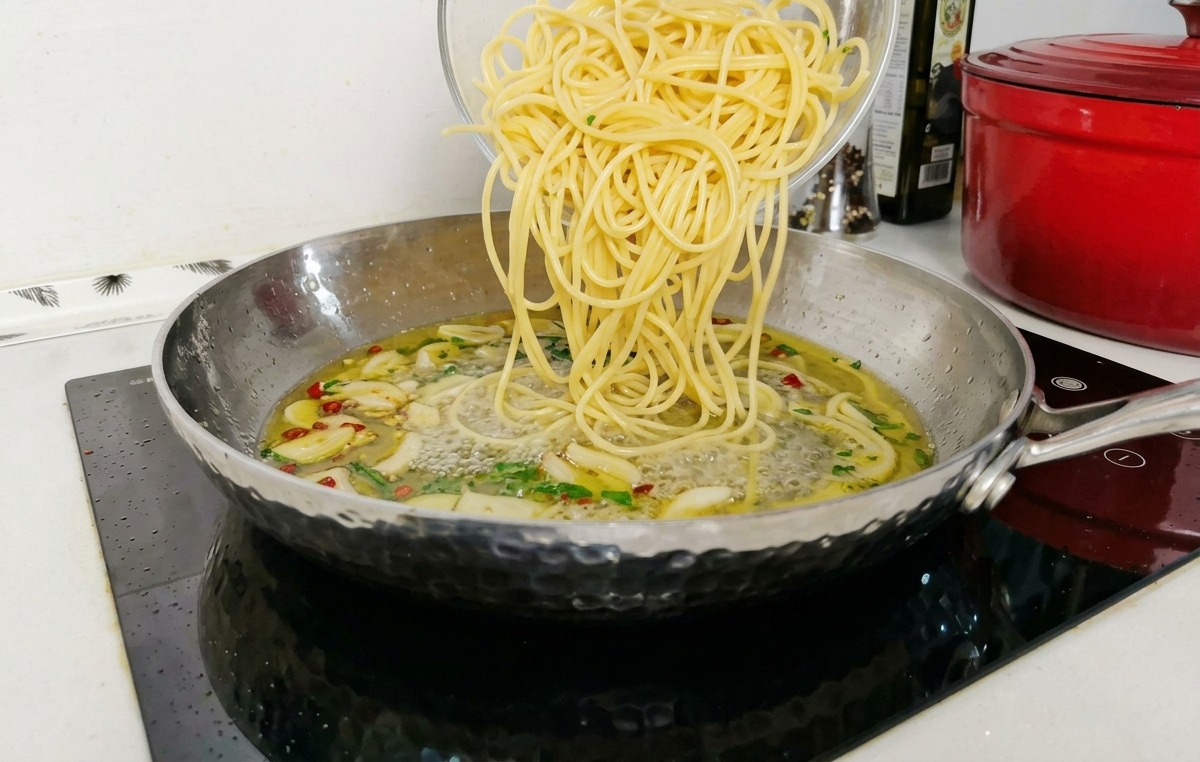 Ugotowane spaghetti przekładane ze szklanej miski na patelnię wypełnioną lekko bulgoczącym sosem czosnkowo-oliwnym.