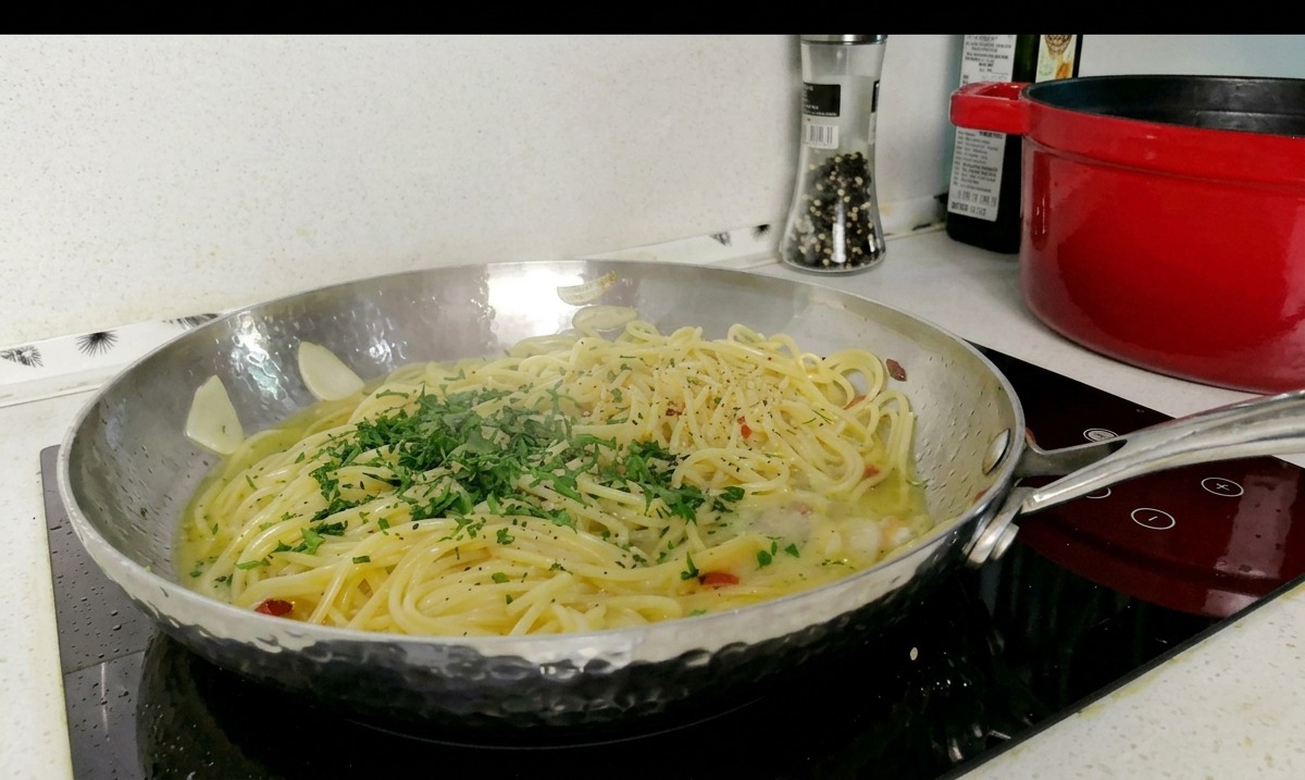 Patelnia ze stali nierdzewnej wypełniona spaghetti w jasnym sosie, posypana sporą garścią świeżo posiekanej natki pietruszki.