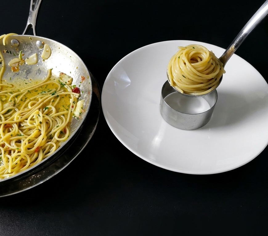 Zbliżenie na spaghetti zawijane za pomocą pęsety w zgrabny cylinder wewnątrz metalowego pierścienia cukierniczego na czystym, białym talerzu.