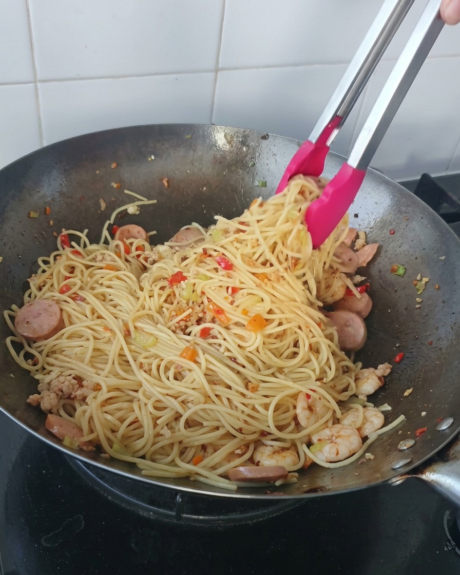 Szczypce z różowymi końcówkami nakładające dużą porcję spaghetti z krewetkami i plasterkami kiełbasy z woka.