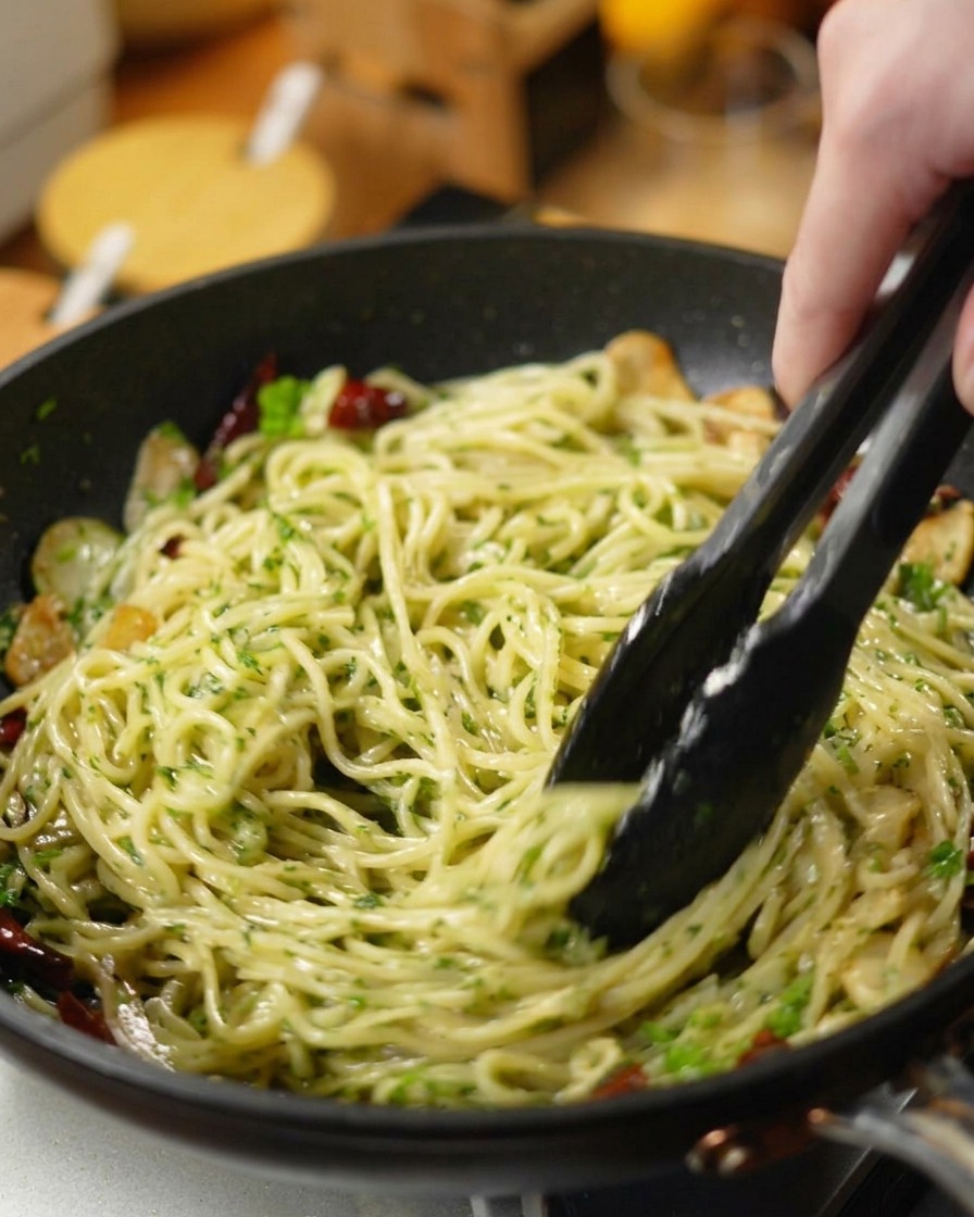 Czarne szczypce mieszające spaghetti z posiekaną pietruszką, czosnkiem i chili na ciemnej patelni.