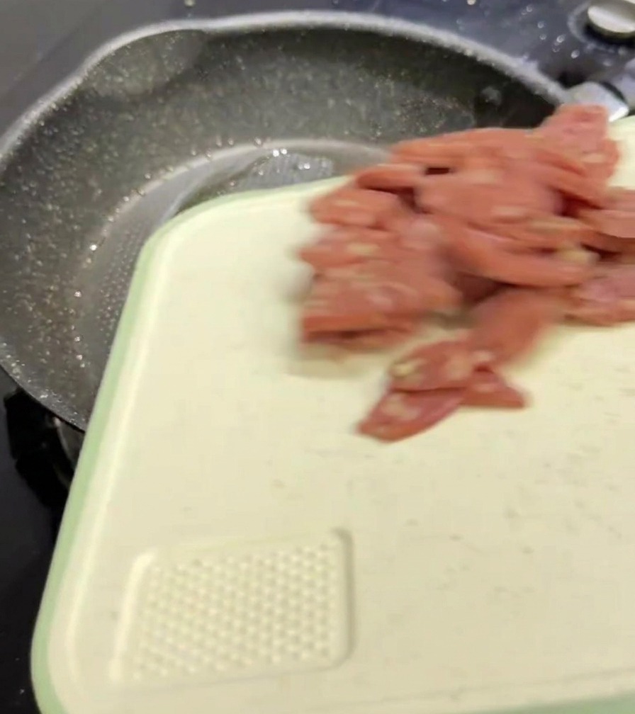 Plasterki różowego salami zsuwające się z białej deski do krojenia na ciemną, nakrapianą patelnię.