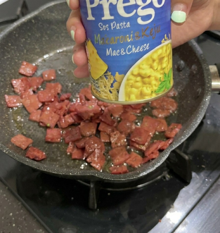 Dłoń trzymająca niebiesko-żółtą puszkę sosu serowego Prego Mac & Cheese nad patelnią z podsmażonymi kawałkami salami.