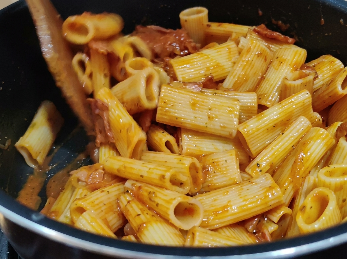 Mieszanie makaronu Rigatoni z gęstym, czerwonym sosem pomidorowym w garnku za pomocą drewnianej łopatki.