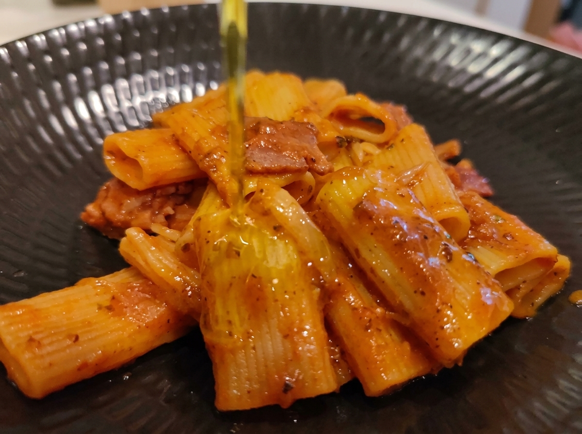 Strumień oliwy z oliwek extra virgin wlewany na nałożoną na talerz porcję rigatoni Amatriciana.