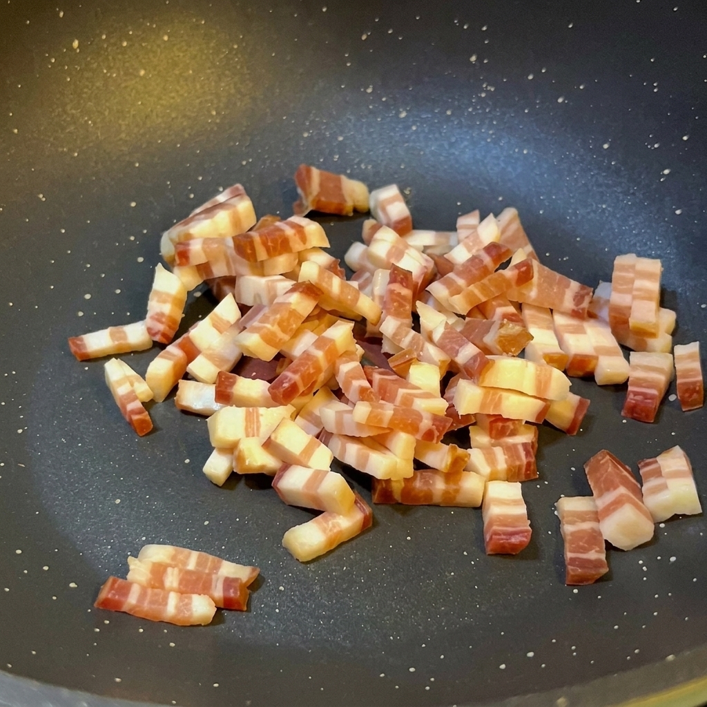 Małe kostki włoskiej pancetty smażą się na ciemnej patelni z nieprzywierającą powłoką, widoczny jest wytopiony tłuszcz i lekkie zrumienienie na brzegach.