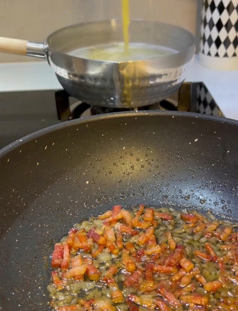 Dłoń używająca metalowych szczypiec do wyciągania żółtego spaghetti z garnka z wrzącą wodą i przekładania go na patelnię z podsmażoną pancettą i cebulą.