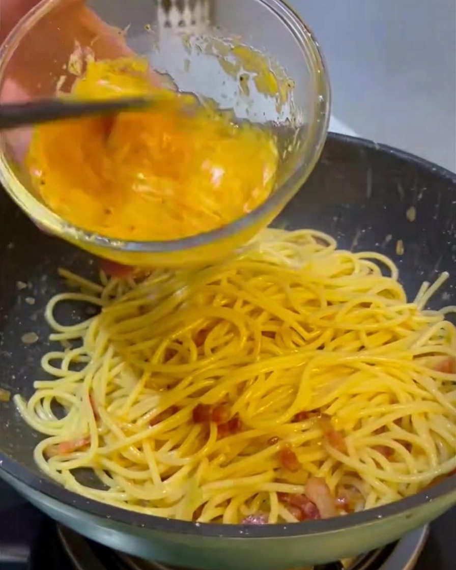 Wlewanie intensywnie żółtej masy z jajek i sera ze szklanej miski na patelnię z gorącym spaghetti i podsmażonym mięsem.