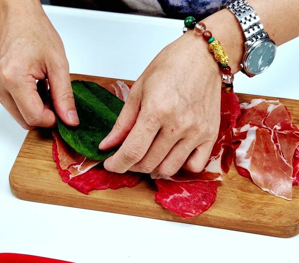 Dłonie kładące duży, świeży, zielony liść szałwii na plastrze cielęciny przykrytym szynką prosciutto.