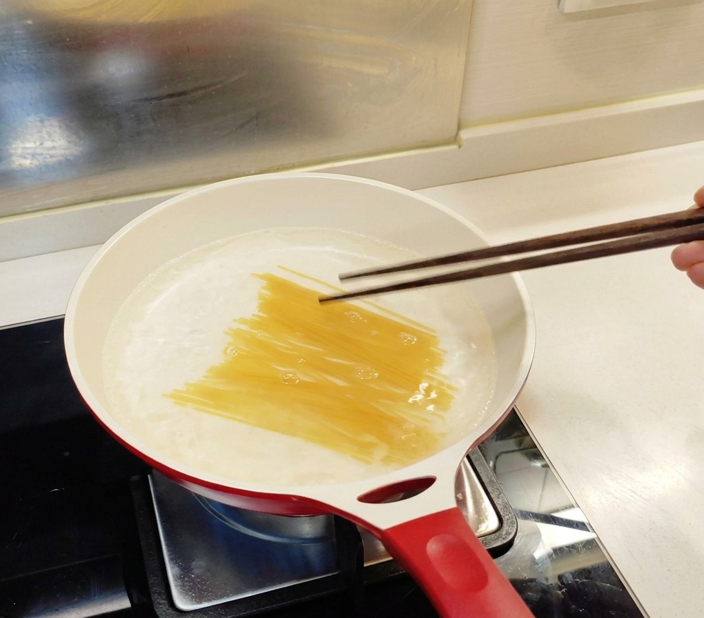 Surowy makaron spaghetti wkładany za pomocą ciemnych drewnianych pałeczek do białego garnka z wrzącą wodą.