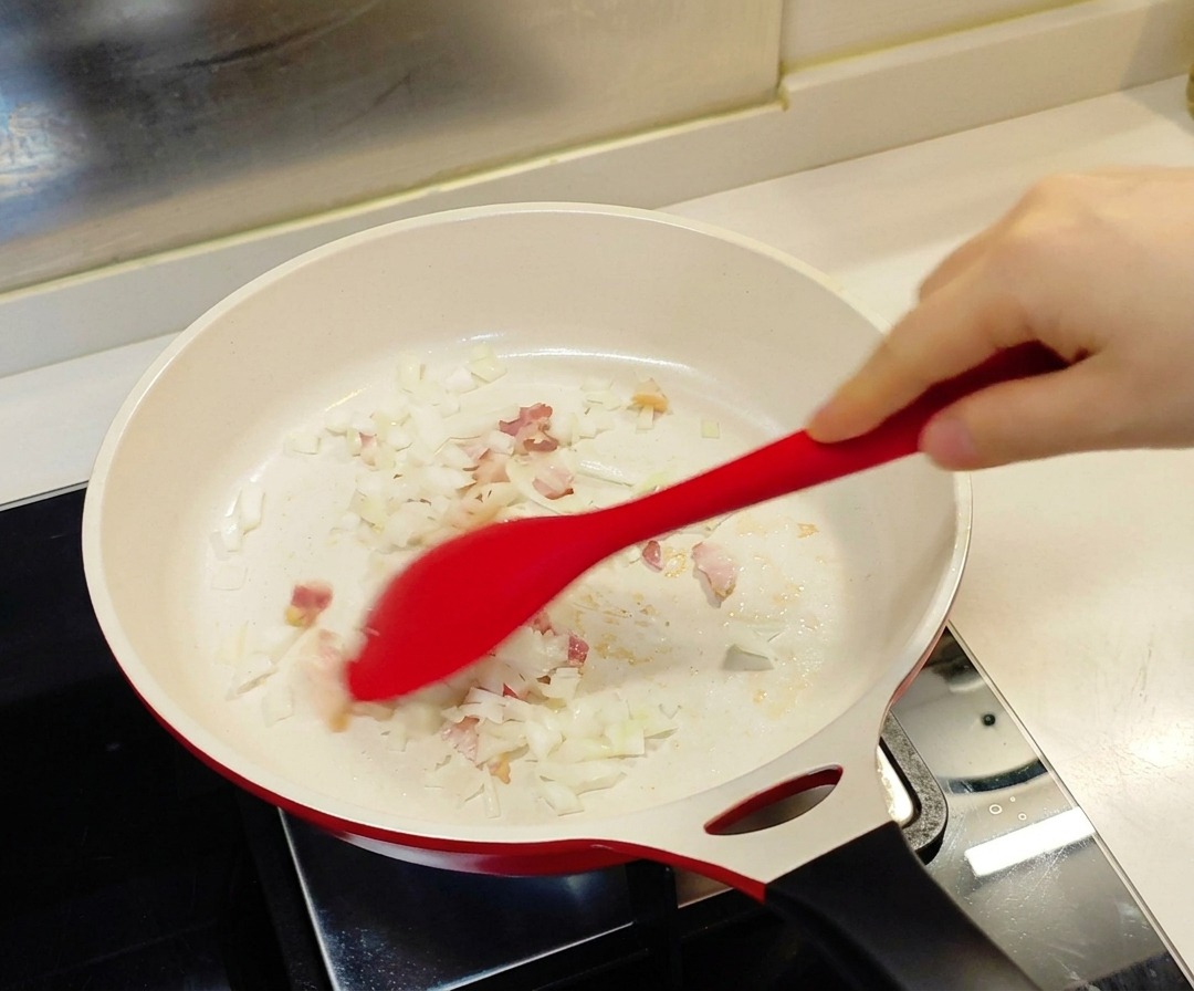 Czerwona łopatka mieszająca pokrojoną cebulę i drobne kawałki boczku na białej patelni z powłoką ceramiczną.