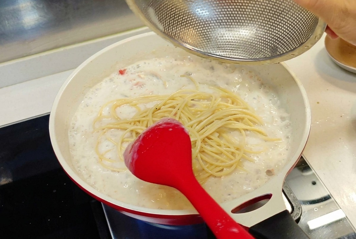 Ugotowany makaron spaghetti przekładany z sitka na patelnię z kremowym białym sosem.