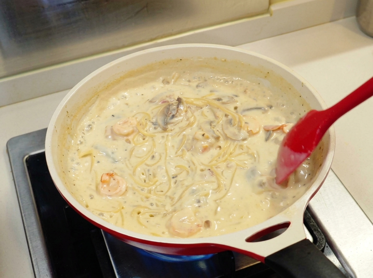 Czerwona łopatka mieszająca spaghetti w gęstym, kremowym białym sosie z krewetkami i pieczarkami na patelni.