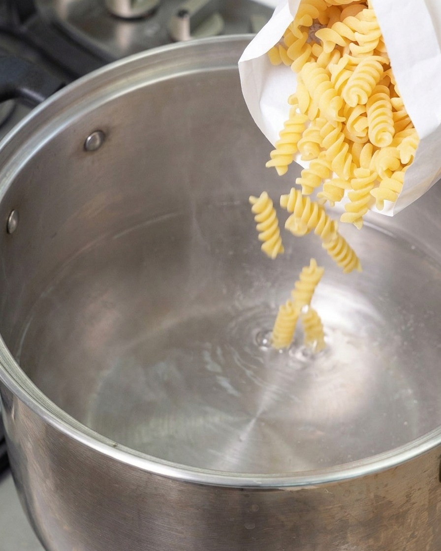 Suchy makaron fusilli wsypywany z białej torebki do dużego garnka ze stali nierdzewnej z wrzącą wodą.