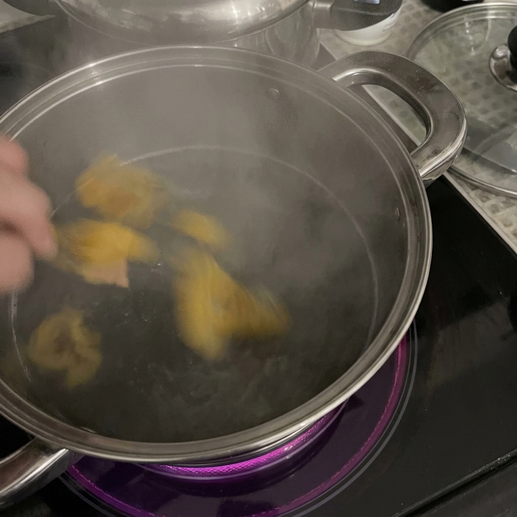 Dłoń wrzucająca żółty makaron rigatoni do garnka ze stali nierdzewnej z parującą, wrzącą wodą na płycie indukcyjnej.