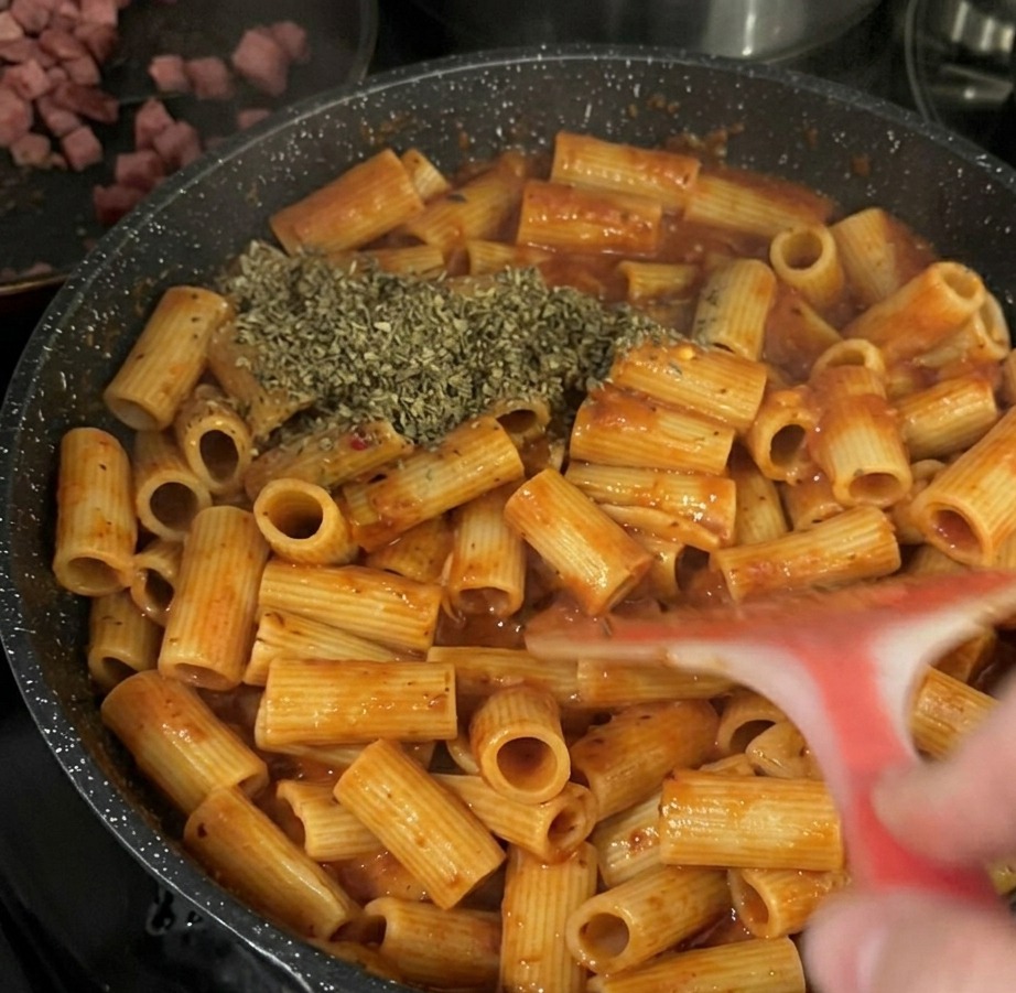 Duża porcja suszonych zielonych płatków bazylii rozsypana na makaronie rigatoni w sosie pomidorowym na czarnej patelni.