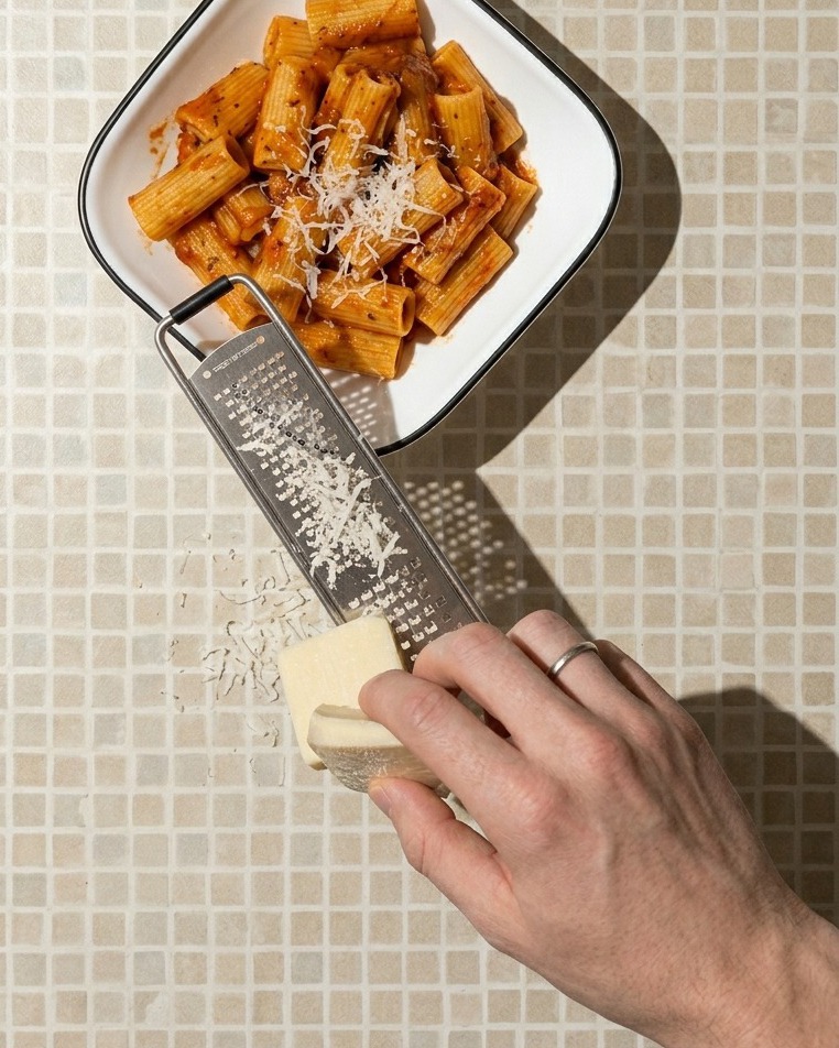 Zbliżenie na dłoń trącą kawałek białego sera nad białą kwadratową miską pełną makaronu rigatoni w czerwonym sosie.