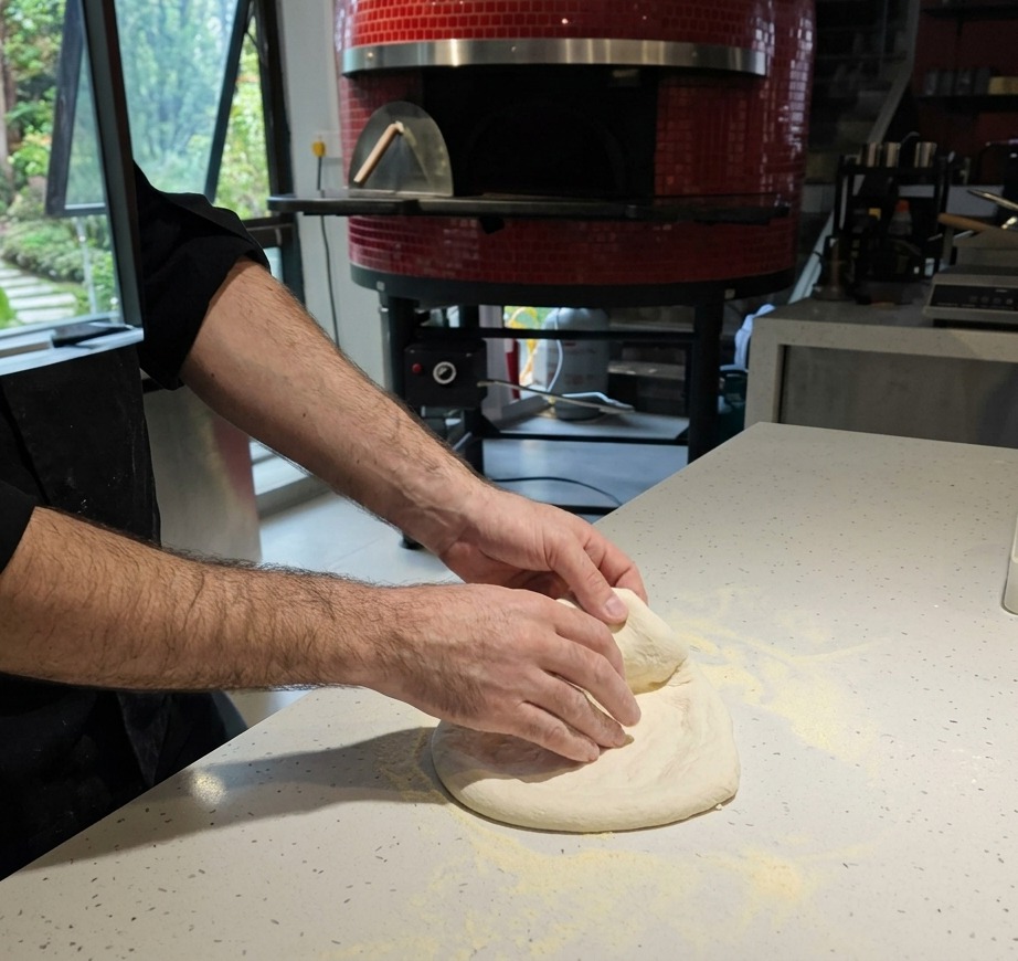 Zbliżenie na dłonie rozciągające surowe ciasto na pizzę na posypanym mąką białym blacie.