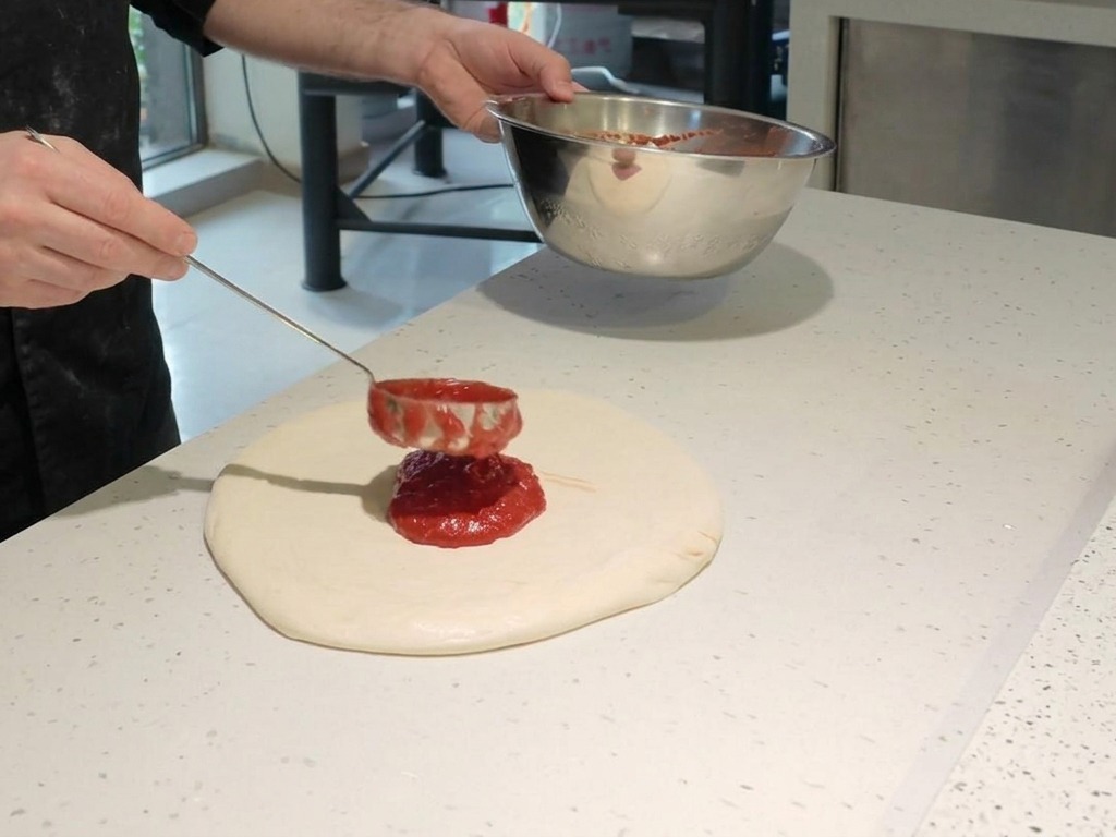 Szef kuchni nakładający metalową chochlą żywoczerwony sos pomidorowy spiralnym ruchem na surowe ciasto na pizzę.