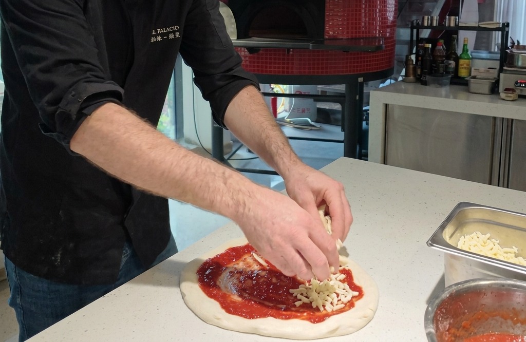 Dłonie układające kawałki białej, świeżej mozzarelli na okrągłym spodzie do pizzy pokrytym czerwonym sosem pomidorowym.