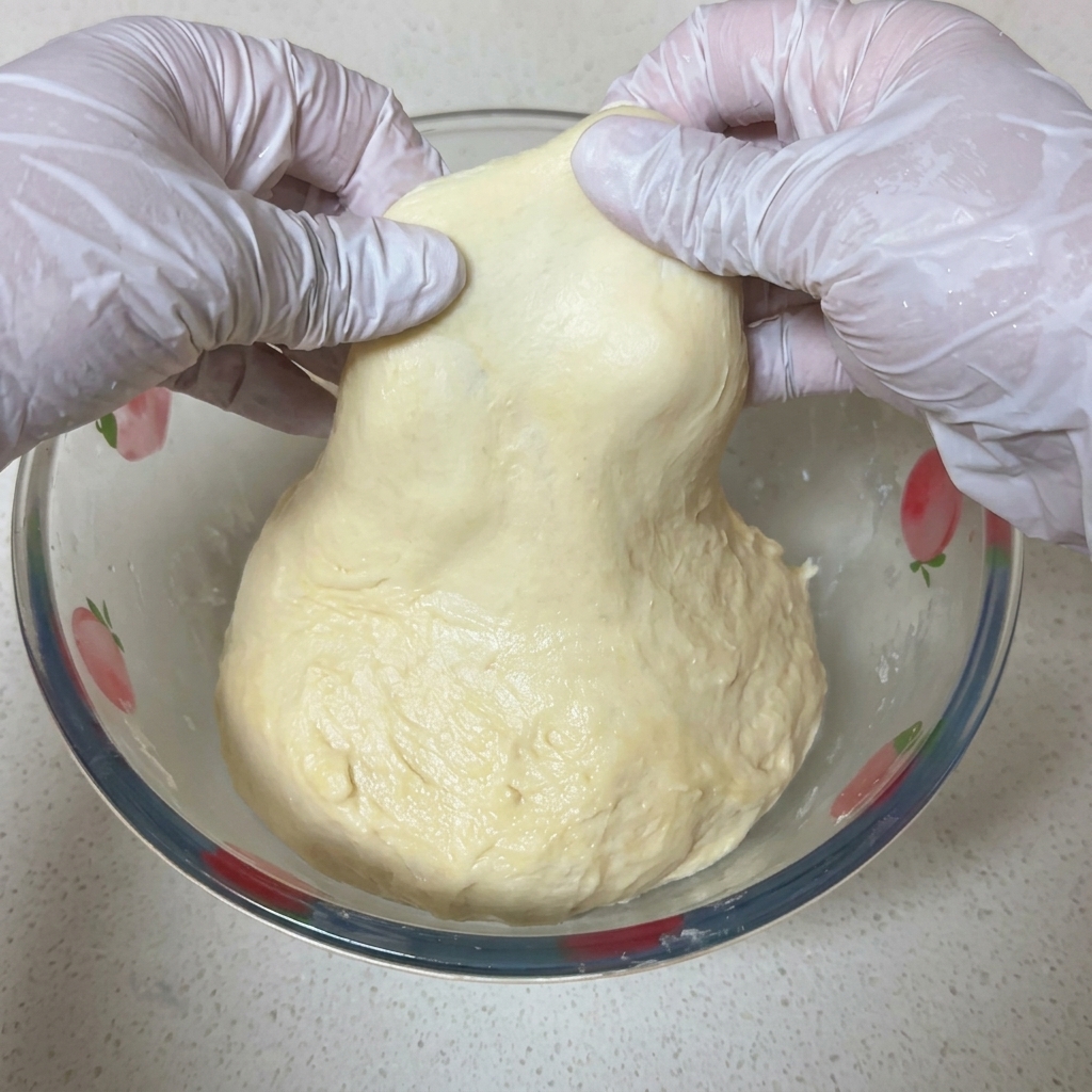 Dwie dłonie w białych rękawiczkach ostrożnie rozciągają kawałek jasnego ciasta na pizzę, pokazując cienką, półprzezroczystą membranę pod światło.