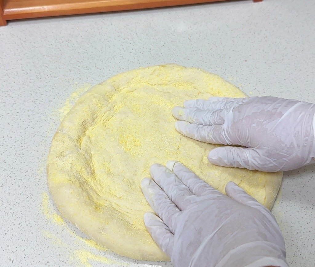 Dłonie w białych rękawiczkach dociskają okrągły kawałek ciasta na pizzę, oprószony żółtą mąką kukurydzianą, na jasnym blacie.