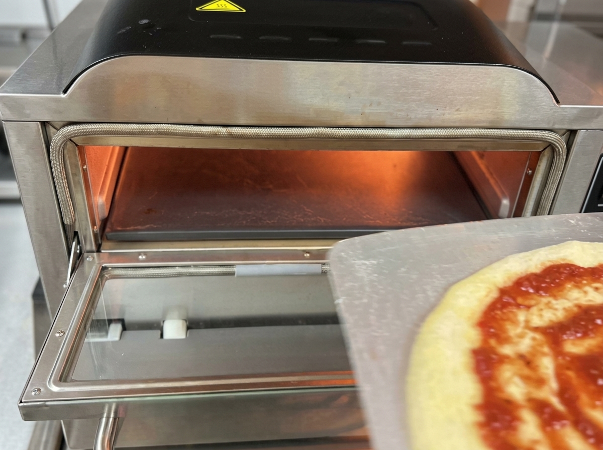 Surowa pizza z sosem i mozzarellą jest zsuwana z metalowej łopaty do rozgrzanego wnętrza profesjonalnego elektrycznego pieca do pizzy.