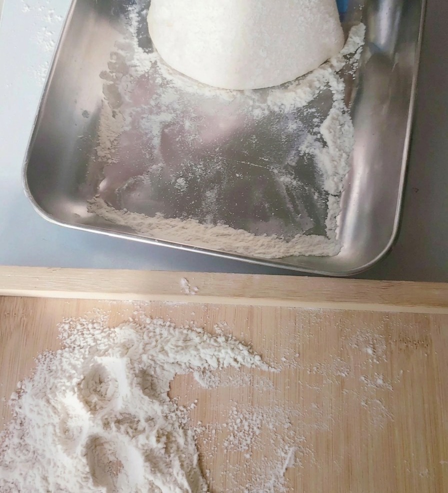 Jasna kulka ciasta na pizzę przenoszona z metalowej tacy na posypaną mąką drewnianą deskę.