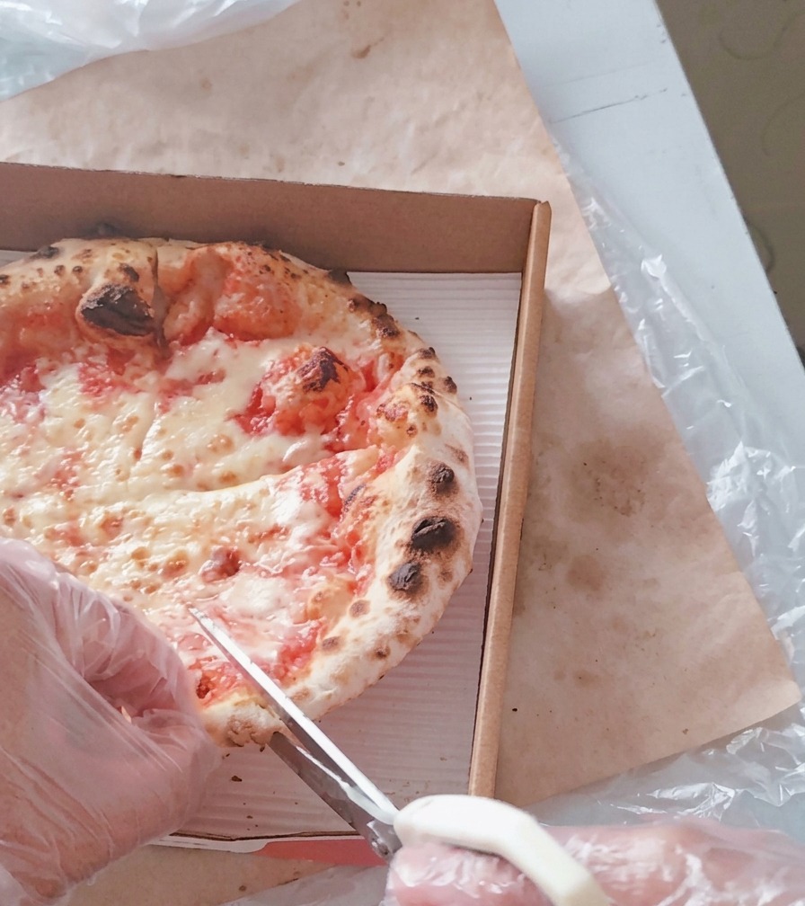 Osoba w rękawiczkach używająca białych nożyczek kuchennych do krojenia gorącej pizzy z serem na trójkąty w kartonowym pudełku.