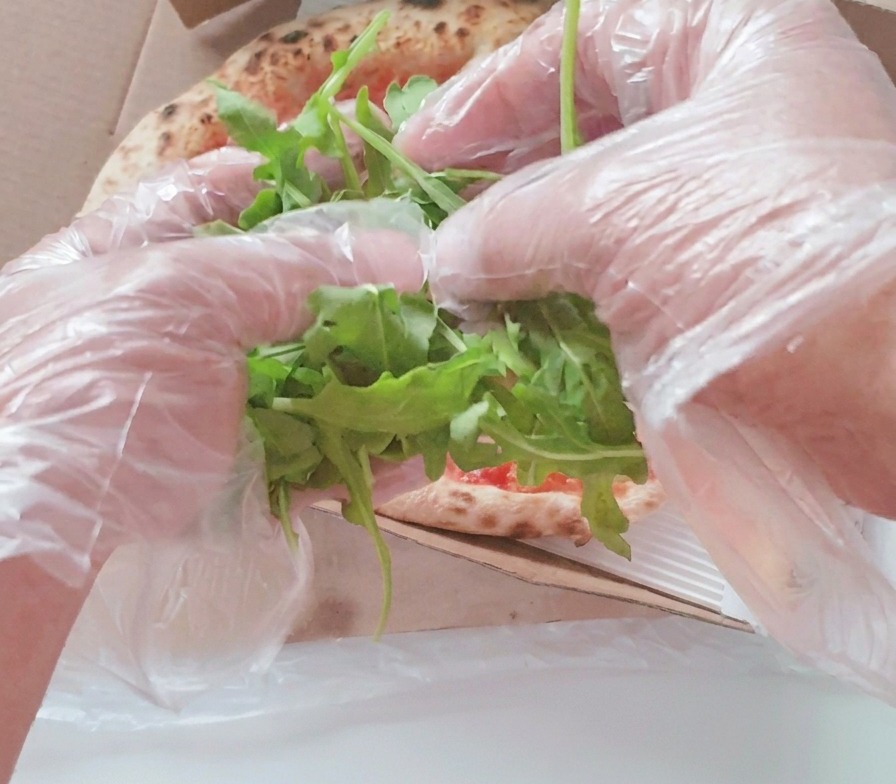 Dłonie w przezroczystych rękawiczkach układające kopczyk świeżych liści rukoli na środku upieczonej pizzy w pudełku.