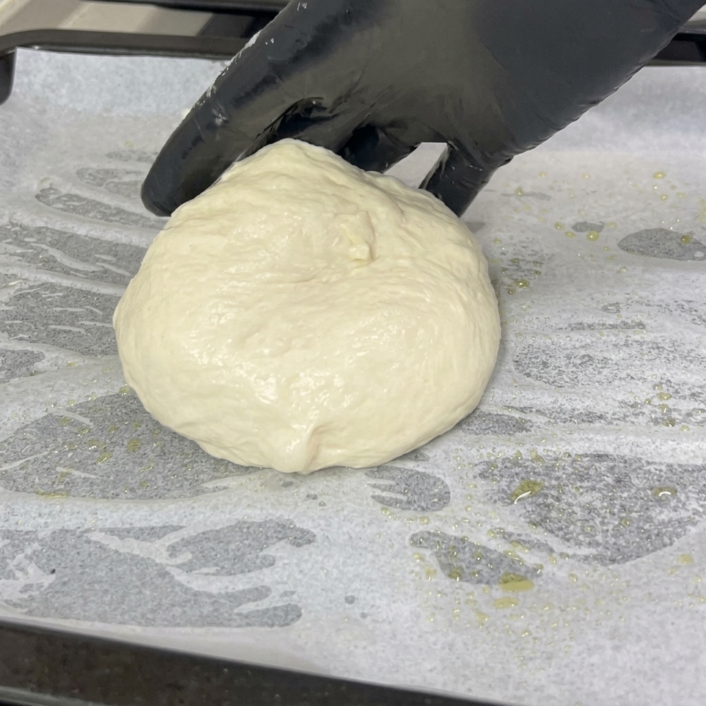 Dłoń w rękawiczce kładzie miękką, zaokrągloną kulę ciasta na pizzę na blachę wyłożoną białym papierem do pieczenia.