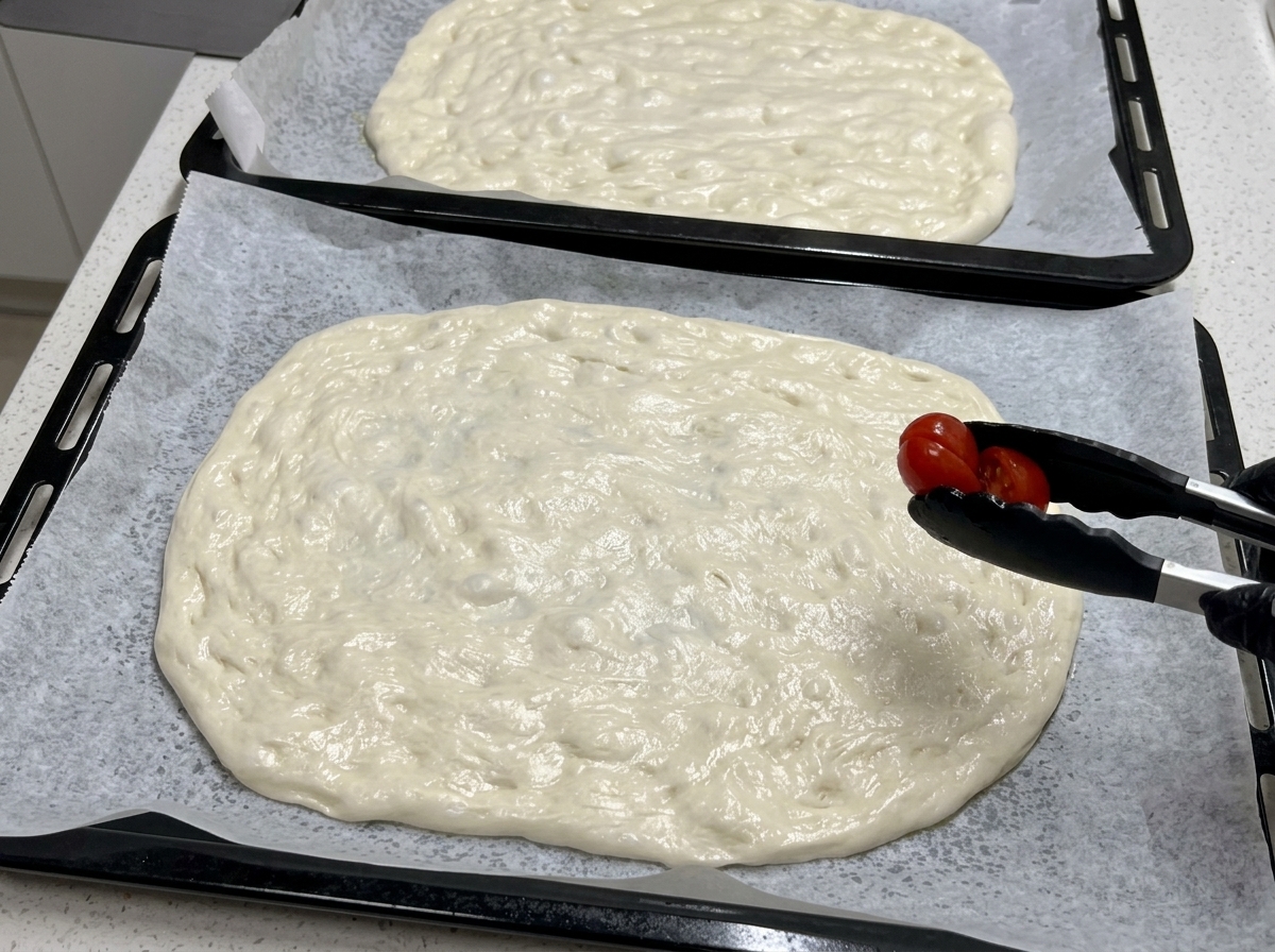 Za pomocą czarnych szczypiec kuchennych połówka pomidorka koktajlowego jest układana na dużym płacie surowego ciasta na pizzę.