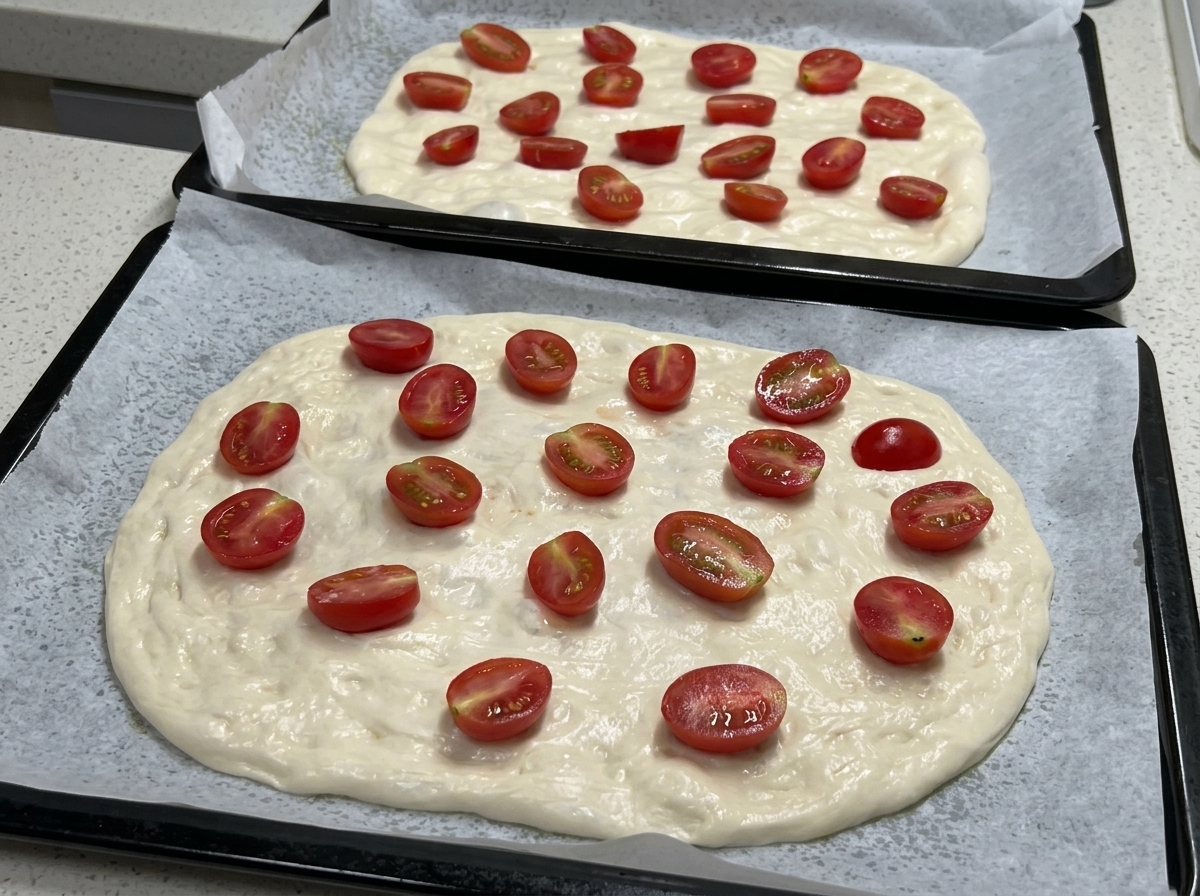 Dwie blachy do pieczenia z rozciągniętym ciastem na pizzę, obficie pokrytym połówkami pomidorków koktajlowych, przygotowane do doprawienia.