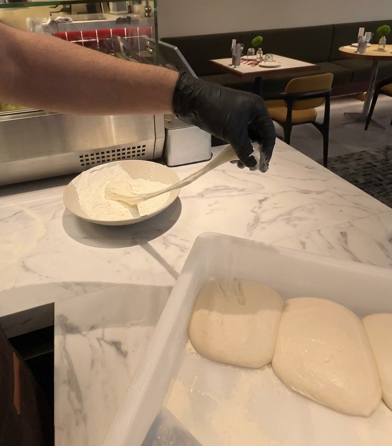 Dłoń w rękawiczce wyjmująca elastyczny kawałek sfermentowanego ciasta na pizzę z miski z białą mąką.