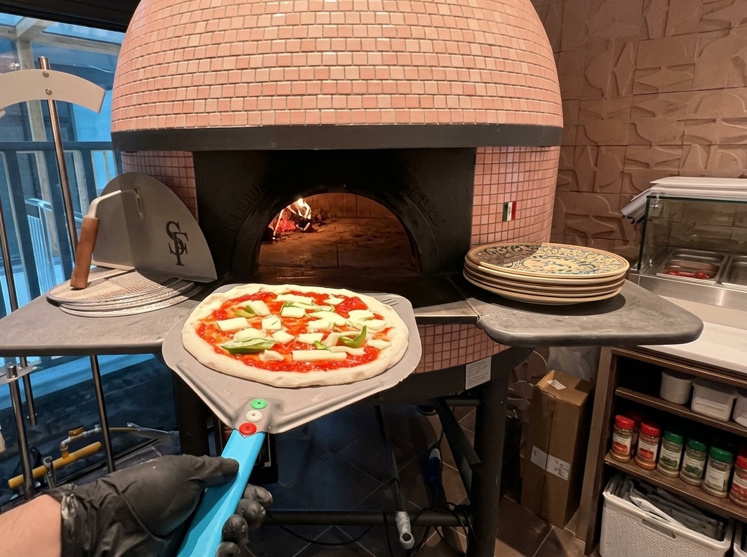Surowa pizza z sosem pomidorowym i mozzarellą na metalowej łopacie przy wejściu do różowego pieca opalanego drewnem.
