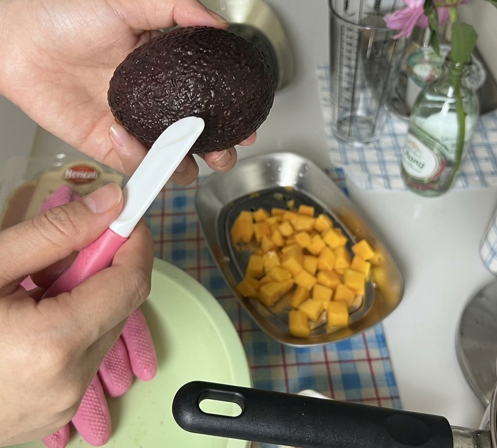 Dłonie krojące obraną połówkę awokado w małą kostkę nad taca z pokrojonym mango.
