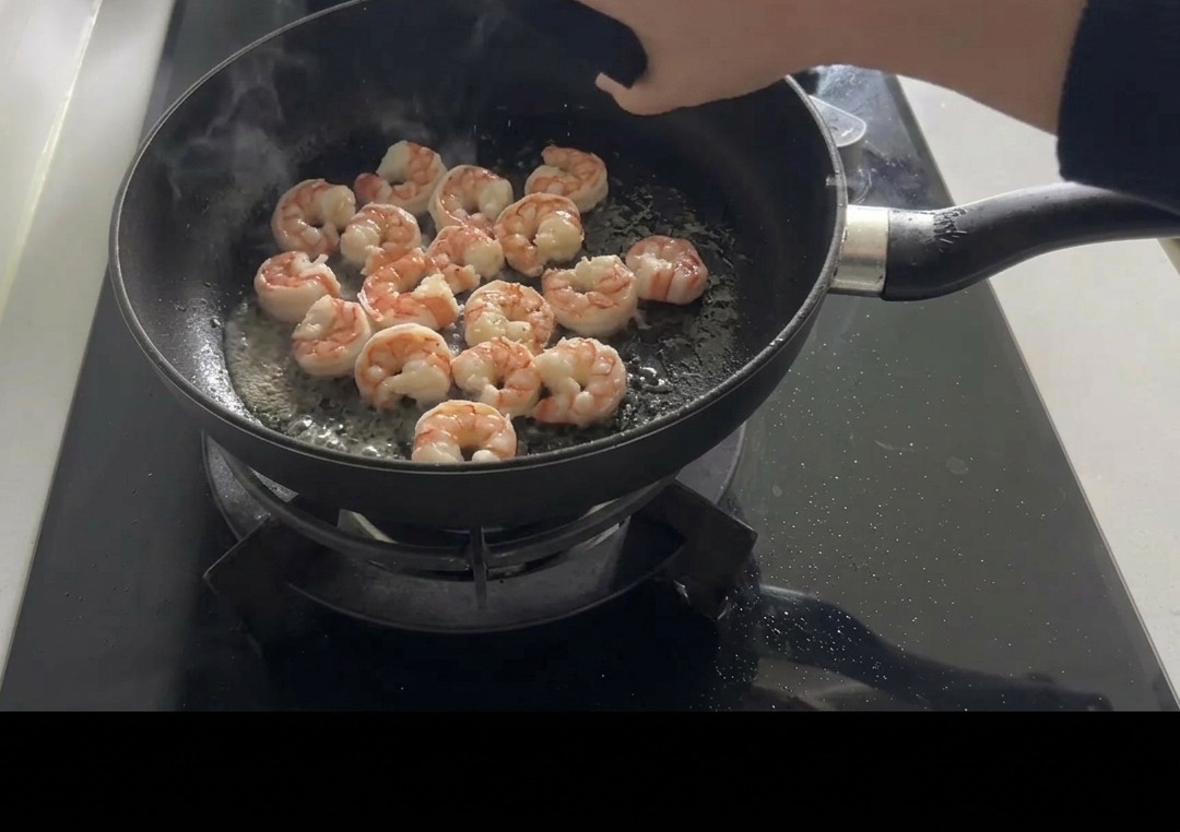 Różowe, usmażone krewetki na czarnej patelni doprawiane ręcznie solą i pieprzem.