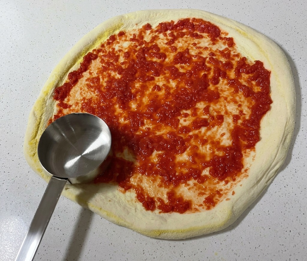 Metalowa chochelka rozprowadzająca krwistoczerwony sos pomidorowy kolistymi ruchami po powierzchni surowego ciasta na pizzę.