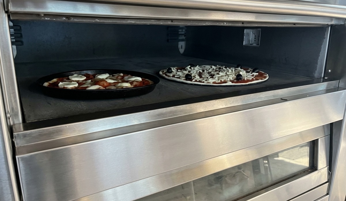 Dwie pizze piekące się na kamiennej powierzchni wewnątrz profesjonalnego, stalowego pieca modułowego.