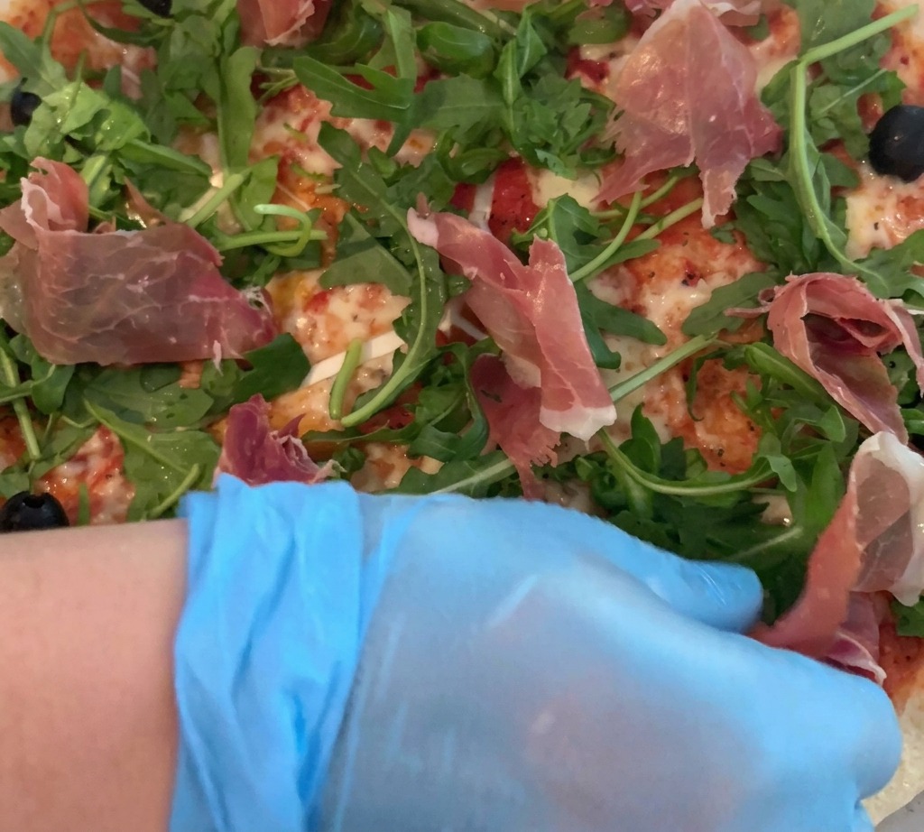Dłoń w rękawiczce ostrożnie układająca cienkie plastry prosciutto na pizzy z rukolą.