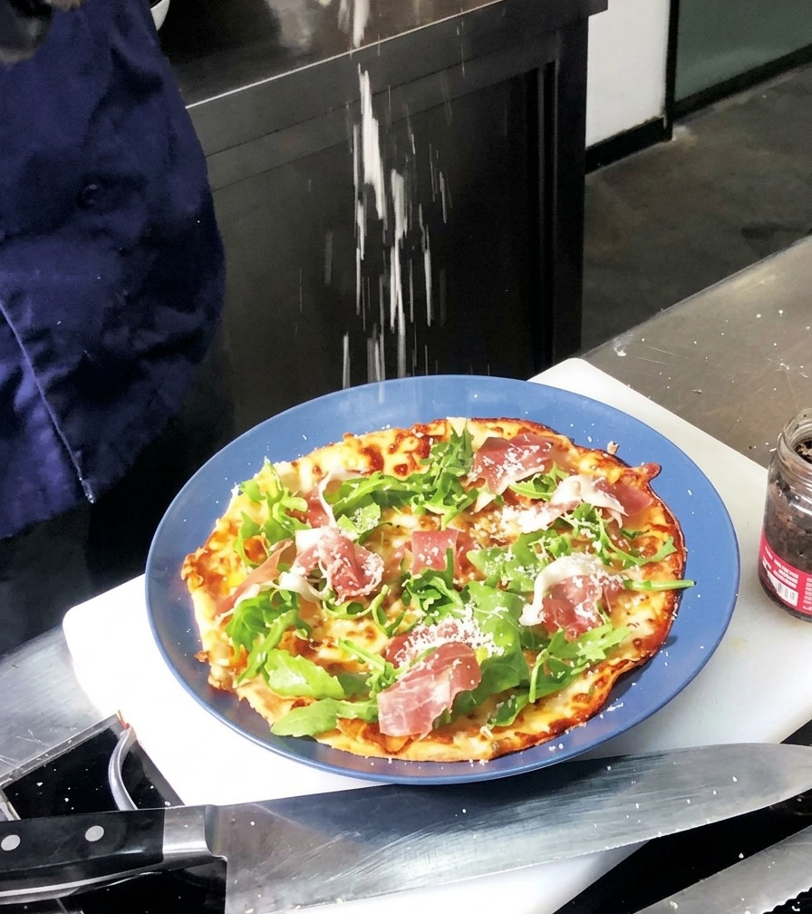 Drobno tarty parmezan sypany na upieczoną pizzę udekorowaną zieloną rukolą i czerwonymi plastrami szynki.