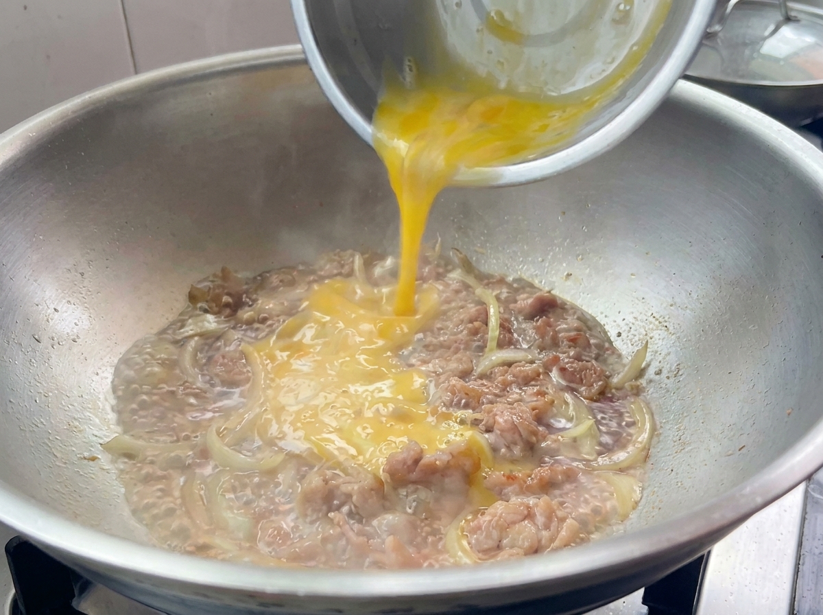 Roztrzepane jajka wylewane na wieprzowinę z cebulą na patelni.